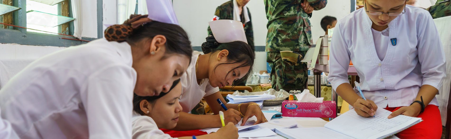 Nurses sit documenting at a table while military staff stand nearby