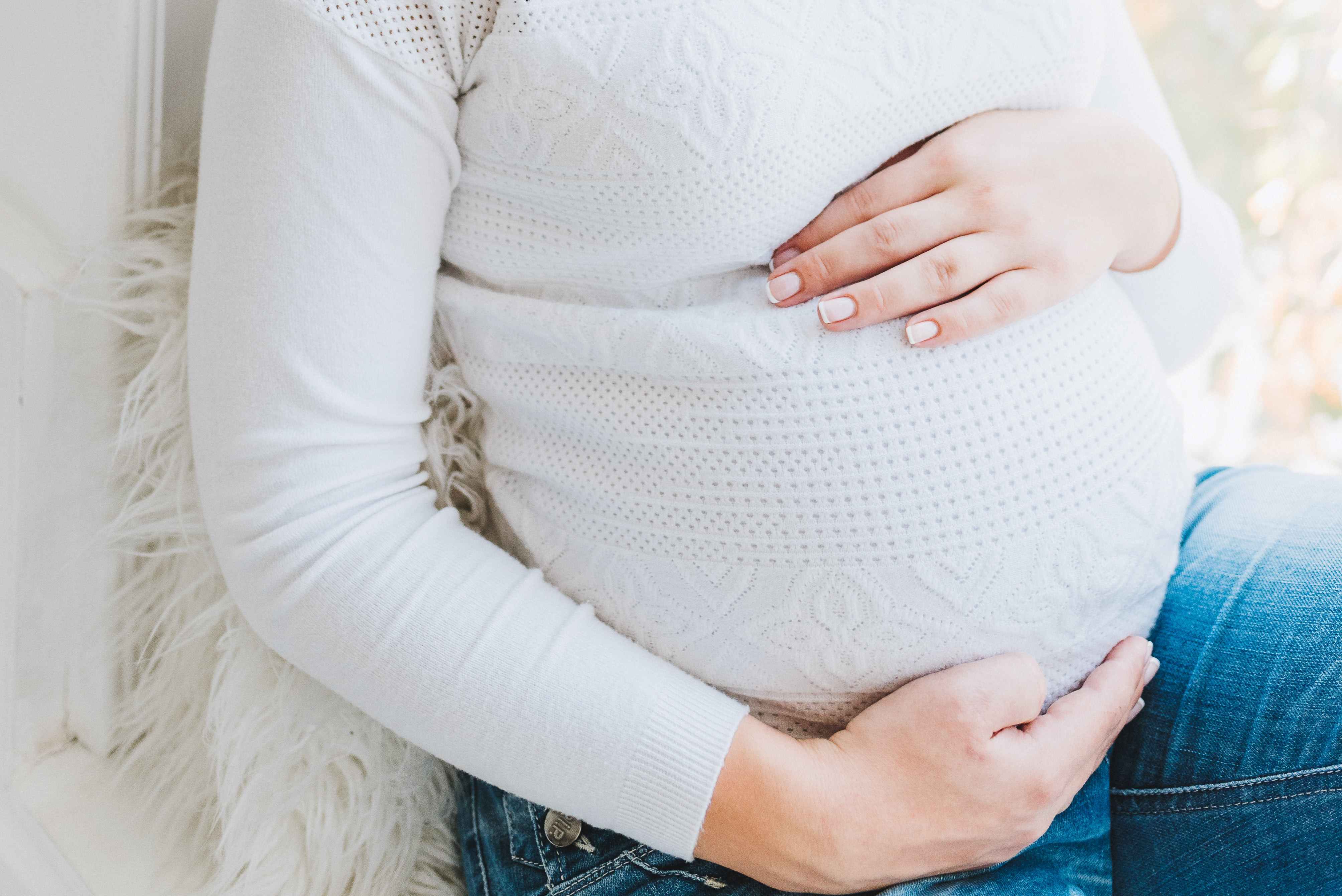 Pregnant woman wearing white is seated on chair covered in white throw