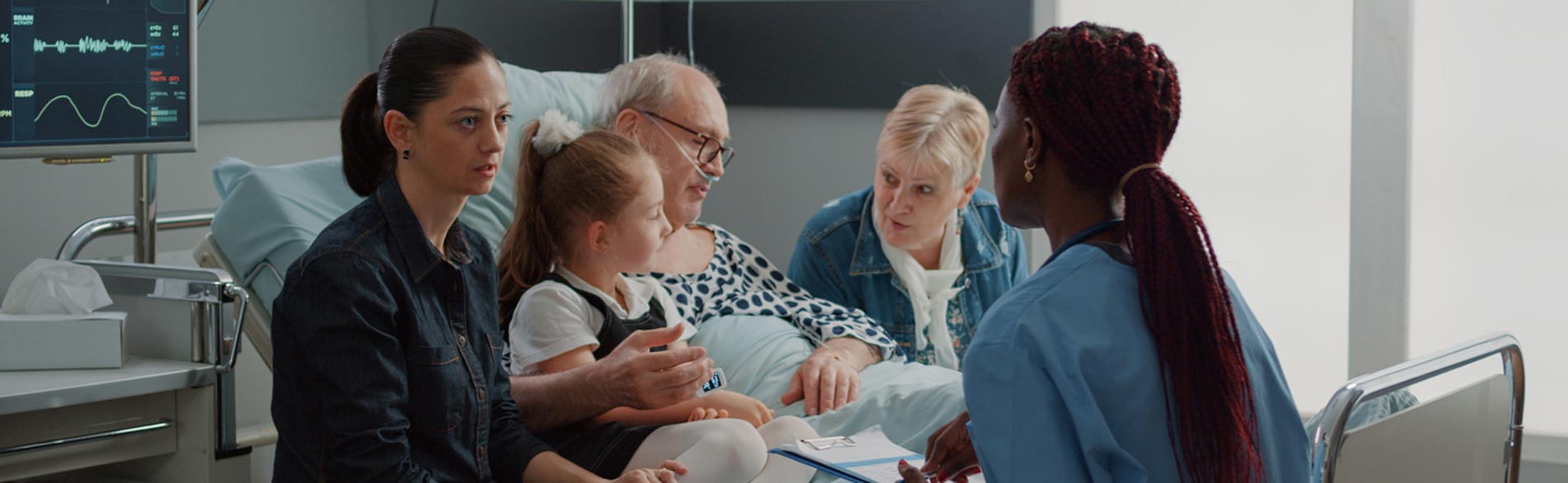 The family of a patient sit around his hospital bed while talking to a clinician.