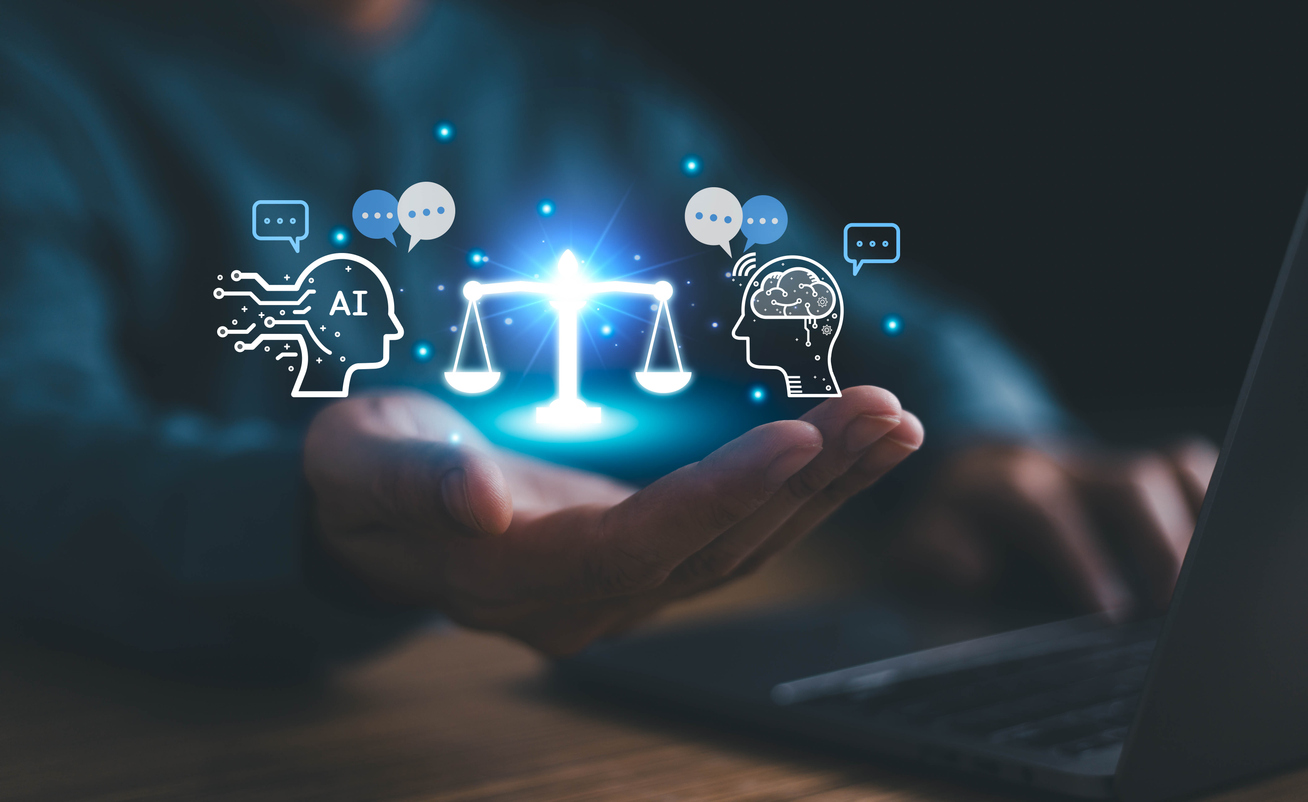 A person’s hand held above a laptop keyboard with digital graphics superimposed. The graphics include a glowing scale symbol in the center, a head icon labeled “AI” on the left with circuit lines, and a human head icon on the right with a brain illustration. Speech bubble icons appear around the heads, representing communication or decision-making between artificial intelligence and human thinking. The scene suggests balance between AI and human judgment.