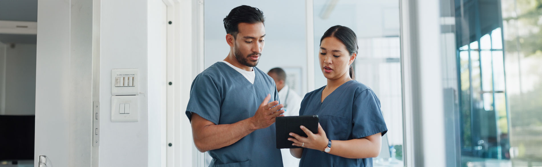 Two healthcare professionals in blue scrubs stand in a bright hospital hallway, discussing something on a digital tablet, with another clinician blurred in the background.