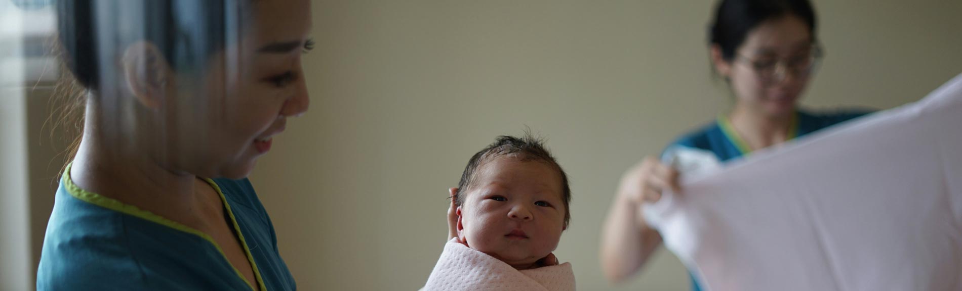 Two midwives in scrubs gently care for a newborn baby wrapped in a white blanket in a softly lit room.