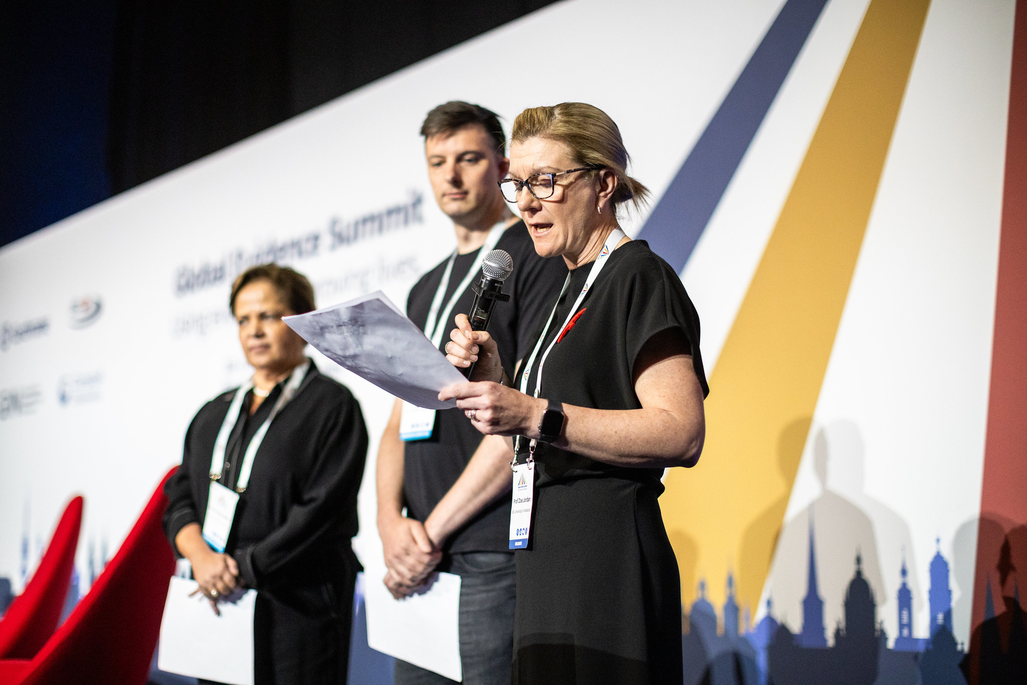 Prof Zoe Jordan announces the beginning of a partnership between JBI, Cochrane and Campbell Collaboration on stage at the Global Evidence Summit. Prof Jordan holds a microphone, and standing to the left of her areDr Karla Soare-Weiss, Cochrane, and Will Moy, Campbell Collaboration